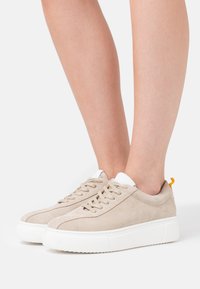 Beiga mockakängor med vita gummisulor, med rund tå, platta snören och en liten gul accent på hälen.