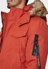 Orange isolierte Jacke mit einer Kapuze aus Kunstpelz, mehreren Taschen, einem schwarzen Reißverschluss und einem Stoffetikett mit "THREADBARE"-Logo.