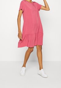 Rosa Kurzarmkleid mit Rundhalsausschnitt und gestuftem Rock. Der Stoff ist glatt und leicht, kombiniert mit weißen Sneakern.