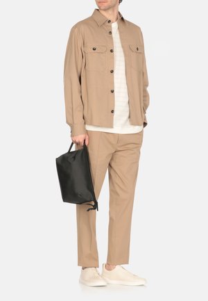 Camicia beige a maniche lunghe con chiusura a bottoni e due tasche sul petto, abbinata a pantaloni coordinati, scarpe chiare e una borsa nera.