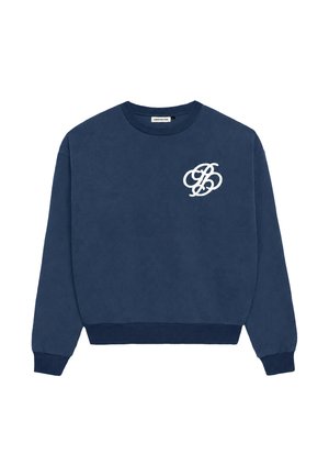 Jonge vrouw met paarse gebreide muts, marineblauwe trui met wit logo, blauwe sjaal met patroon om haar nek en lichtgrijze broek met handen in de zakken.