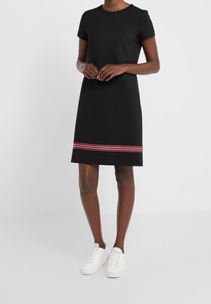Femme debout portant une robe noire mi-longue à manches courtes avec une bande rayée rouge et blanche près de l'ourlet, associée à des baskets blanches.