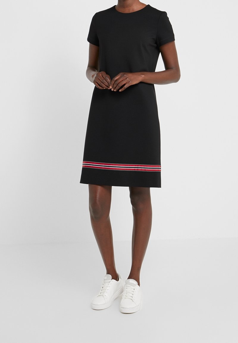 Femme debout portant une robe noire mi-longue à manches courtes avec une bande rayée rouge et blanche près de l'ourlet, associée à des baskets blanches.