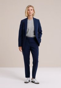 Jūras spēku pielāgots uzvalks ar šauru piegriezumu, apvienots ar gaiši pelēku džemperi un baltām sporta apaviem. Gluda auduma virsma un tīras līnijas visā apģērbā.