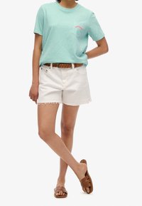 T-shirt di cotone azzurro chiaro con logo a tasca rossa, abbinata a pantaloni denim bianchi con orli sfrangiati. Cintura intrecciata marrone e sandali marroni.