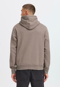 Young man in a light brown hoodie, viewed from behind, standing against a plain white background. Dark pants are visible.