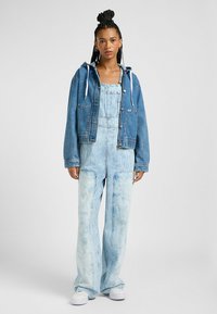 Farkkutakki hupulla ja etutaskuilla, yhdistettyinä vaaleansinisille, leveille haalareille, joissa on pystysuorat raidat ja haalistu väri.