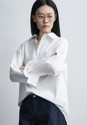 Jeune femme aux cheveux noirs lisses portant des lunettes transparentes, une chemise blanche ample boutonnée, un collier de perles et un pantalon foncé, les bras croisés.