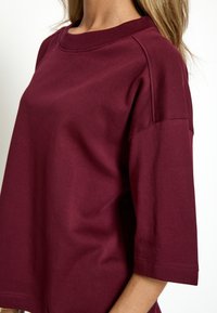 T-shirt oversize burdeos con cuello redondo, mangas cortas, cuello de canalé y una textura suave de algodón. Presenta un corte holgado y un diseño sencillo.