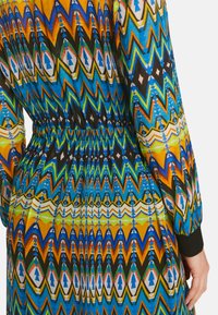 Mehrfarbiges Kleid mit einem Zickzackmuster in Blau, Gelb und Grün. Elastischer Taillenbund und schwarz abgesteppte Ärmel. Weicher, leichter Stoff.