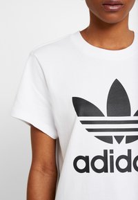 Bílý bavlněný tričko s černým logem Adidas trefoil a textem. Krátké rukávy, kulatý výstřih, hladká textura.