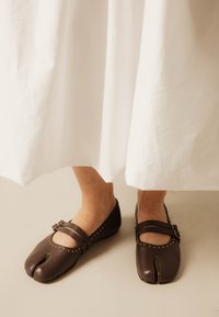 Pieds portant des chaussures Mary Jane marron à bout fendu avec des clous dorés, visibles sous une longue jupe blanche.