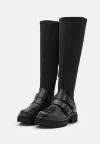 Svarta knähöga stövlar med strukturerat läder, dubbla spännen och en gummisula med mönstrat mönster. Dragkedja på sidan.