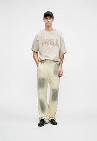 T-shirt bej oversized cu inscriptiona "FAVELA" în bej mai închis, asociat cu pantaloni crem cu stropi întunecați și încălțăminte neagră.