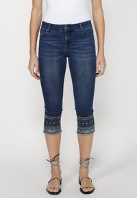 Jean capri en denim bleu foncé avec ourlet embellit. Présente un design ajusté, des poches avant et un petit bord retroussé. Détails texturés sur les poignets.