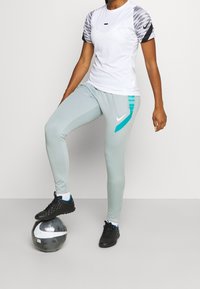 Nike Sportoutfit bestehend aus einem weißen kurzärmeligen Oberteil mit grauem Muster auf den Ärmeln, hellgrauen Leggings mit türkisfarbenen Akzenten und schwarzen Sportschuhen.