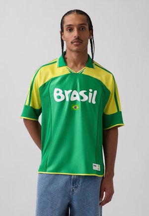 Muž s pletenými vlasmi nosiaci zelený a žltý brazílsky futbalový dres s textom "Brasil" a vlajkou stojaci pred jednofarebným pozadím.