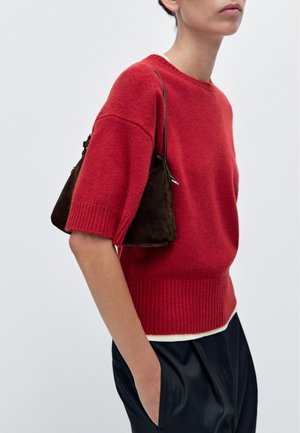 Person wearing a red short-sleeve sweater, black pants, and carrying a small dark brown shoulder bag, with one hand in pocket.