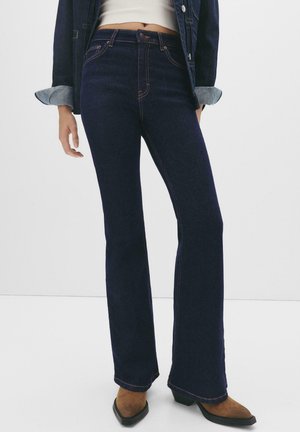 Donkerblauwe flared jeans met contrasterende oranje stiksels, hoge taille en een zachte denimtextuur. Gepaard met bruine puntige laarzen.