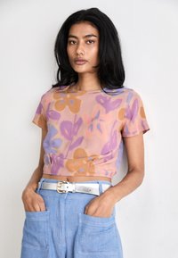 Short-sleeve, cropped blouse in a sheer fabric with a pastel floral pattern in purple, orange, and pink. Paired with light blue pants and silver belt.