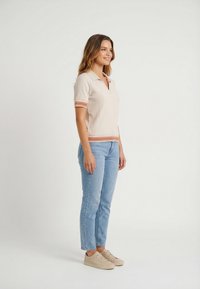 Mujer sonriente de pie, vistiendo un suéter de manga corta con cuello color beige claro, jeans azul claro y zapatillas beige, sobre un fondo liso.