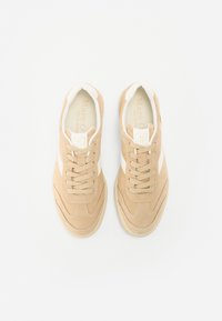 Beige velúrpóló cipők fehér oldalsávokkal, kerek orral, fűzős kialakítással és tagolt gumitalppal. A márka logója látható a bélésen.