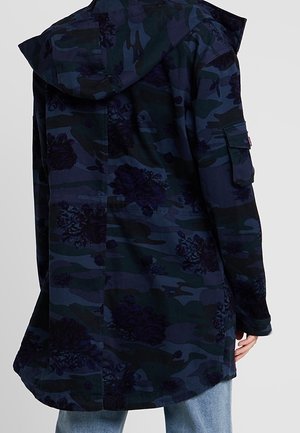 Longue veste camouflage bleu foncé avec motifs floraux, capuche et poche à rabat sur la manche, portée par-dessus un jean bleu clair.