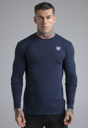 Uomo con tatuaggi che indossa una maglia aderente a maniche lunghe blu navy con logo bianco sul petto, in piedi davanti a uno sfondo grigio uniforme.