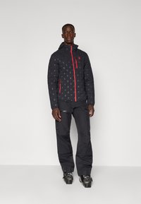 Black puffer jacket with red zipper and logo, featuring a patterned surface. Paired with black pants and sturdy footwear.