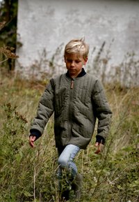 Bambino con giacca trapuntata verde con cerniera, jeans blu e stivali di gomma, che cammina attraverso erba alta e piante.