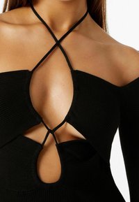 Černý žebrovaný top s hlubokým výstřihem a překříženými ramínky. Materiál je měkký a hladký, což zdůrazňuje strukturovaný design.