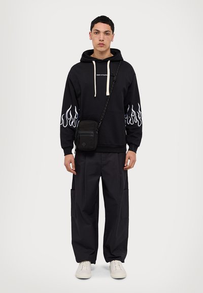 Sudadera negra con gráficos de llamas blancas en las mangas, cordones beige, combinada con pantalones cargo negros y zapatillas crema. Bolsa cruzada pequeña.