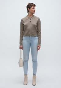 Μπεζ πουλόβερ με γιακά και τρία κουμπιά, συνδυασμένο με ανοιχτόχρωμο στενό τζιν παντελόνι. Το μοντέλο κρατά μια μικρή μπεζ τσάντα, φορώντας ανοιχτόχρωμες άνετες μπότες στον αστράγαλο.