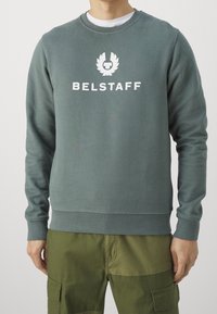 Hombre que lleva un sudadera de microfibra en color verde apagado con el logotipo y el texto de Belstaff en blanco en el pecho, combinada con unos pantalones cargo en color verde oliva.