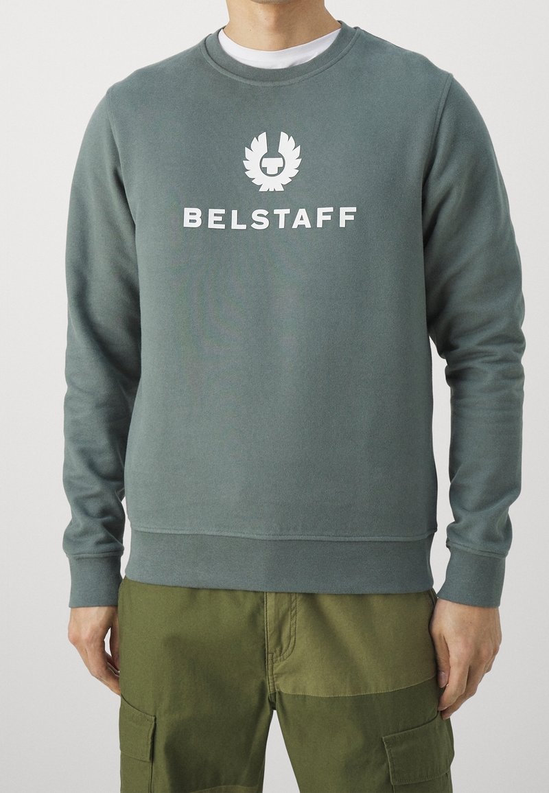 Hombre que lleva un sudadera de microfibra en color verde apagado con el logotipo y el texto de Belstaff en blanco en el pecho, combinada con unos pantalones cargo en color verde oliva.