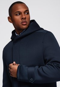 Abrigo capucha azul marino confeccionado con tela estructurada, con un diseño ajustado, detalles de botones en los puños y una textura suave.