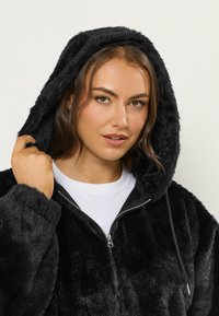 Czarna bluza z sztucznego futra z zamkiem błyskawicznym, kapturem ze sznurkiem i ściągaczami przy rękawach. Miękka tekstura, luźny fason, noszona na białej koszuli.