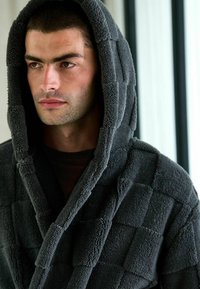 Grå huva av plyschtyg, med ett strukturerat rutmönster. Tyget har en mjuk, lojt utseende som ökar värmen och komforten.