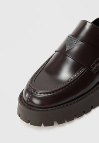 Mocasini din piele lacuită maro, cu talpă groasă din cauciuc negru, având o bandă decorativă și un accesoriu metalic triunghiular pe partea superioară.