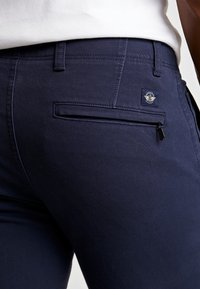 Tmavě modré džíny z bavlny, s zadní kapsou na zip a malou znakem značky. Hladká textura a přiléhavý střih.