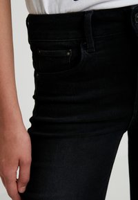 Jeans de mezclilla negra con una cintura alta, bolsillos frontales con tachuelas metálicas y un ajuste ceñido, que destacan la ligera textura de la tela.