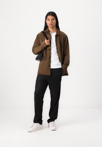 Jachetă din suede maro, cu nasturi, tricou alb, pantaloni negri și adidași albaștri. Geantă mică neagră cu curea. Texturi netede și croială ajustată.