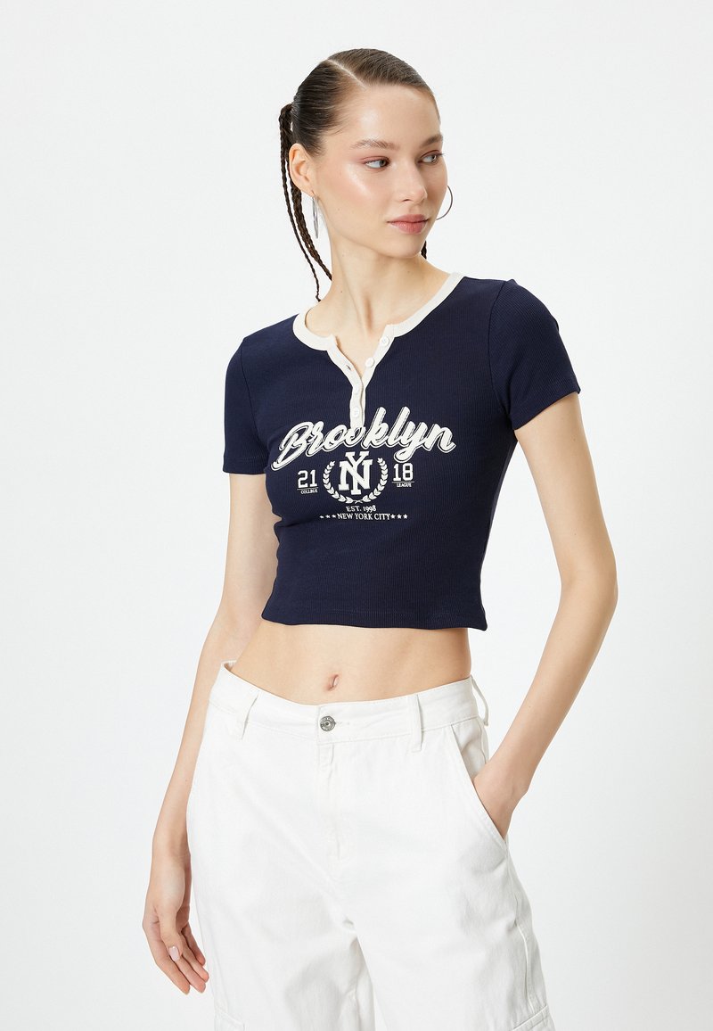 Top corto azul marino con mangas cortas, con una tapeta de botones crema y un gráfico de "Brooklyn" en blanco, combinado con pantalones blancos de cintura alta.