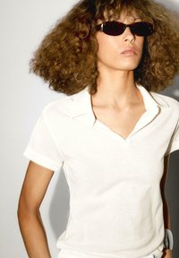 Bílá polo košile s límečkem a krátkými rukávy. Výrazná textura kontrastuje s hladkými slunečními brýlemi. Pozadí je jemně šedé.