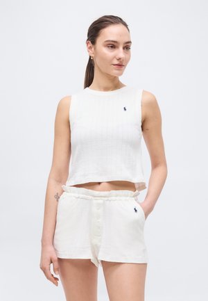 Moteris su kuoduku, vilkinti rankovių neturinčią baltą trumpą palaidinę ir baltus šortus su mažais siuvinėjimo logotipais, stovinti viena ranka kišenėje.