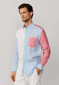 Uomo che indossa una camicia a maniche lunghe con blocchi di colore bianco, azzurro e rosso, in piedi con una mano in tasca.