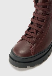 Bottine en cuir bordeaux avec des coutures proéminentes, surface texturée, semelle en caoutchouc noir et lacets plats. Présente une forme arrondie et un design durable.