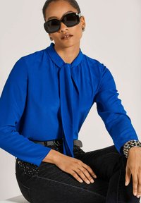 Blaue Bluse mit langen Ärmeln und gebundenem Kragen, kombiniert mit schwarzen Jeans mit Nietenakzenten. Das Model trägt große schwarze Sonnenbrillen.