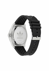 Montre avec un bracelet en silicone noir et un boîtier en acier inoxydable, présentant un dos lisse en argent avec le logo Adidas et des détails texturés sur le bracelet.