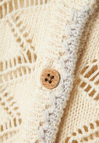 Stickad cardigan i krämfärg med ett öppet vävdesign, prydd med en träknapp med blommönster och en flätad kant i ljusbeige.